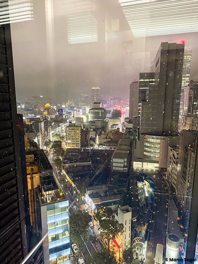 Una veduta notturna di Shibuya presa dall'interno di Cafe Legato con edifici illuminati e pioggia sulla vetrata.