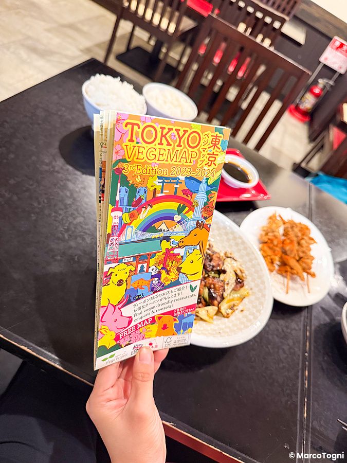 Una mano sostiene una copia della Tokyo Vegemap, mappa con i posti vegani e vegetariani a Tokyo, sopra un tavolo del Saira...