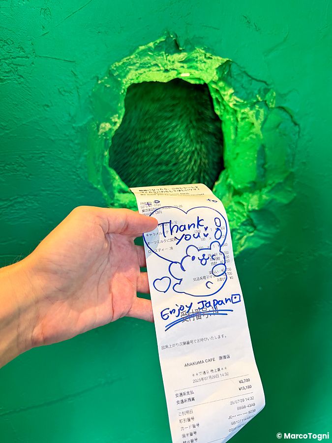 Una mano sostiene uno scontrino dell'Anakuma Cafe davanti al caratteristico foro nel muro utilizzato per il servizio.