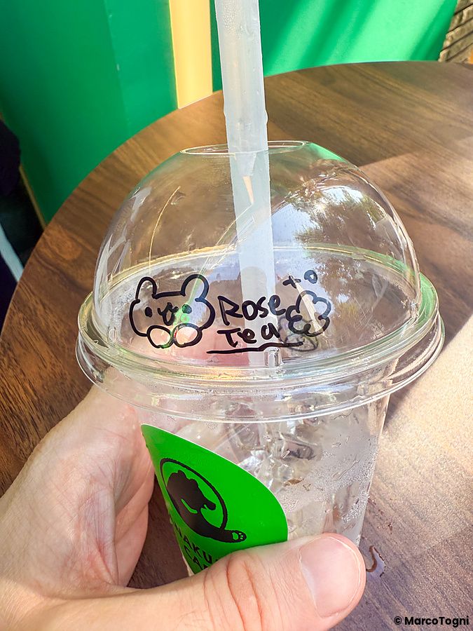 Una mano sostiene un bicchiere di plastica con il logo del Anakuma Cafe e un disegno a mano sul coperchio.