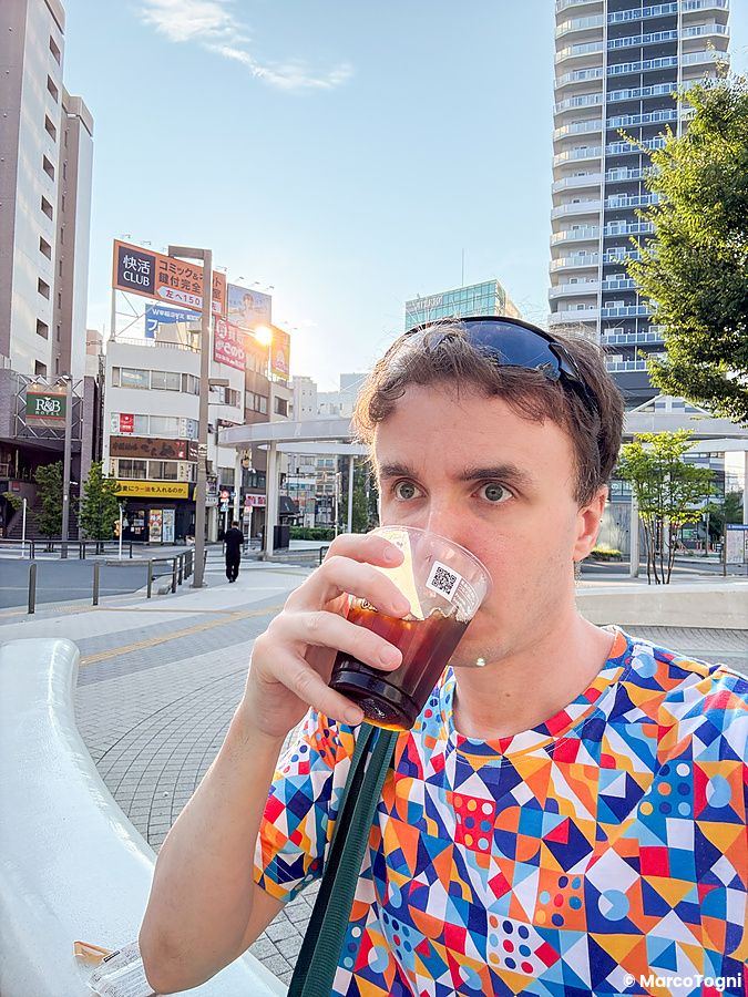 Marco Togni beve un caffè del 7 Eleven ad Otsuka davanti alla stazione, con edifici urbani e cartellonistica sullo sfondo.