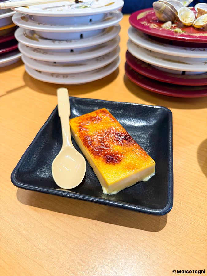 Un dolce alla crema servito su un piatto nero in un ristorante Sushiro, circondato da pile di piatti usati.