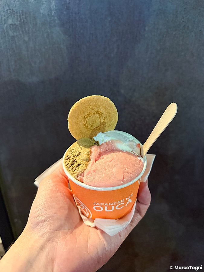 Un gelato in un vasetto arancione con il marchio Ouca tenuto in mano da una persona con un cucchiaino di legno.