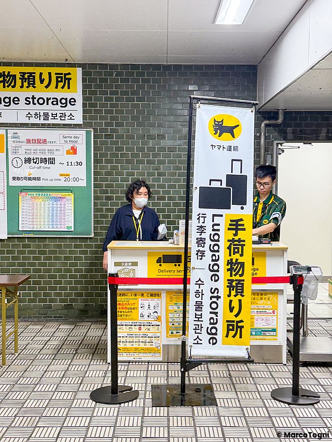 Due operatori lavorano presso un banco di deposito bagagli in stazione in Giappone, segnalato da stendardi gialli e neri.