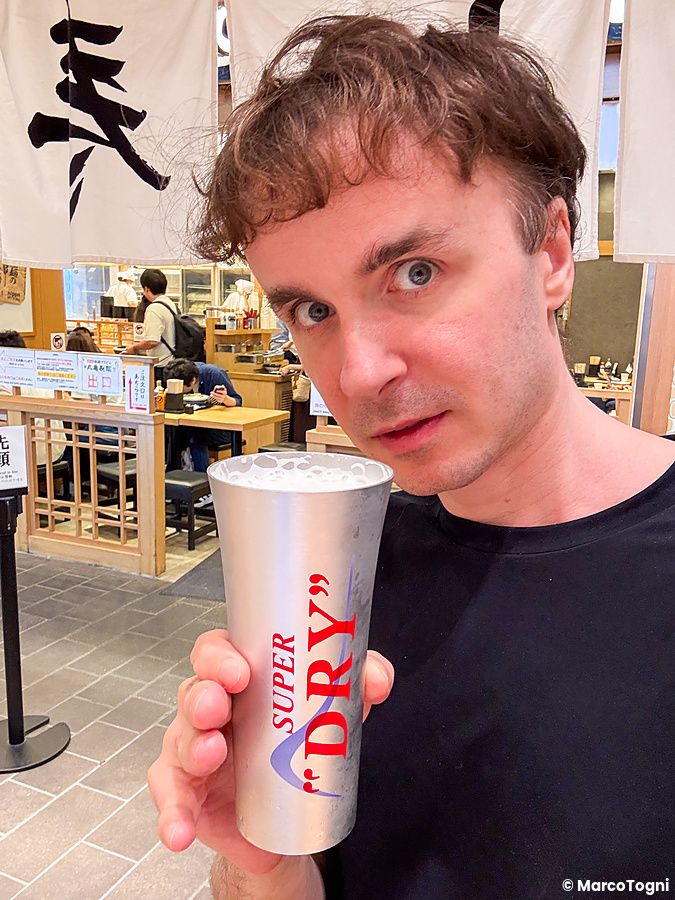 Marco Togni tiene un bicchiere metallico di birra Asahi in un locale in Giappone, con sullo sfondo persone ai tavoli.