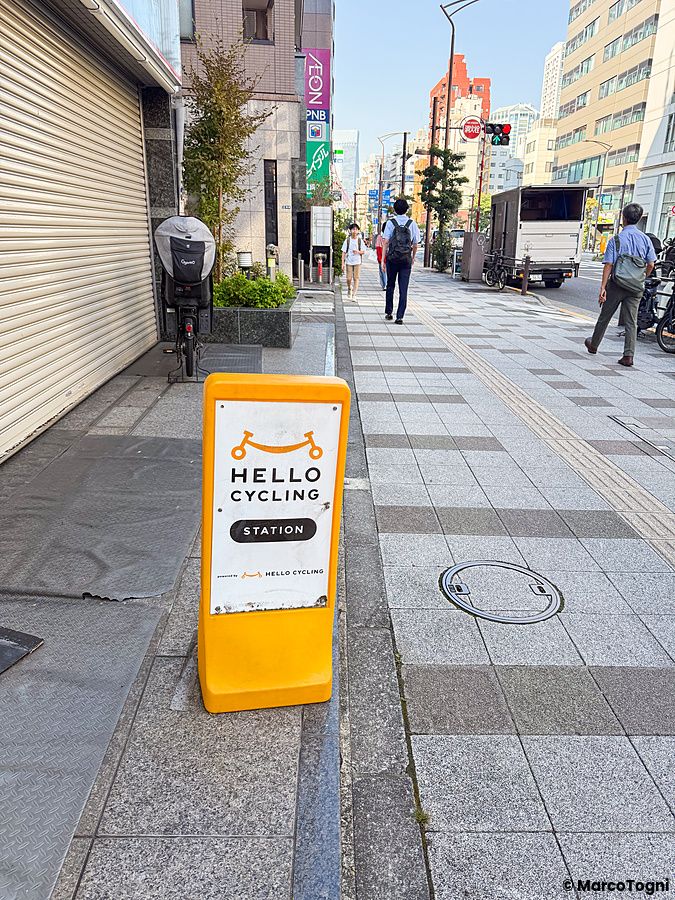 Cartello giallo di una stazione di noleggio bici in Giappone con il logo Hello Cycling su un marciapiede a Tokyo.