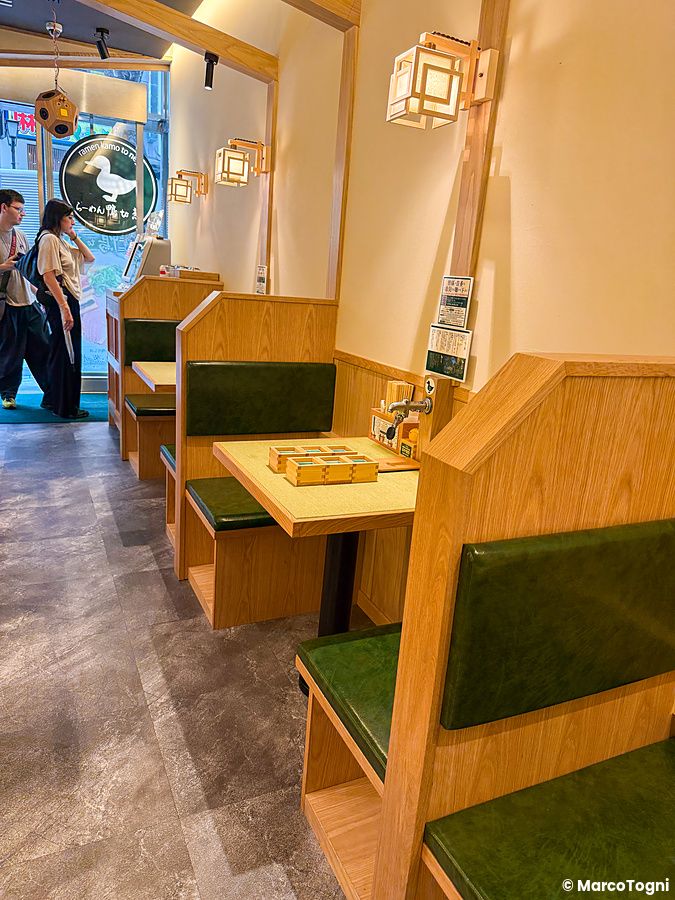Interno del ristorante Ramen Kamo to Negi con tavoli in legno, sedute verdi e lampade a lanterna in Giappone.