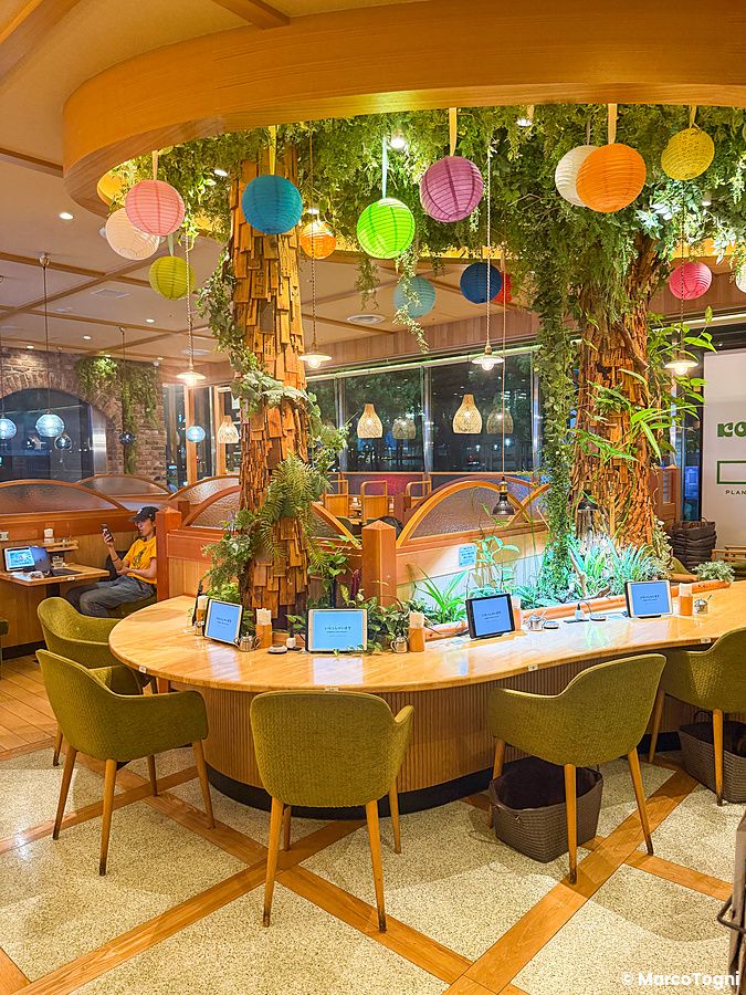 Interno del ristorante vegano Komeda Is a Ginza, con tavoli circondati da piante, lanterne colorate e una persona seduta.