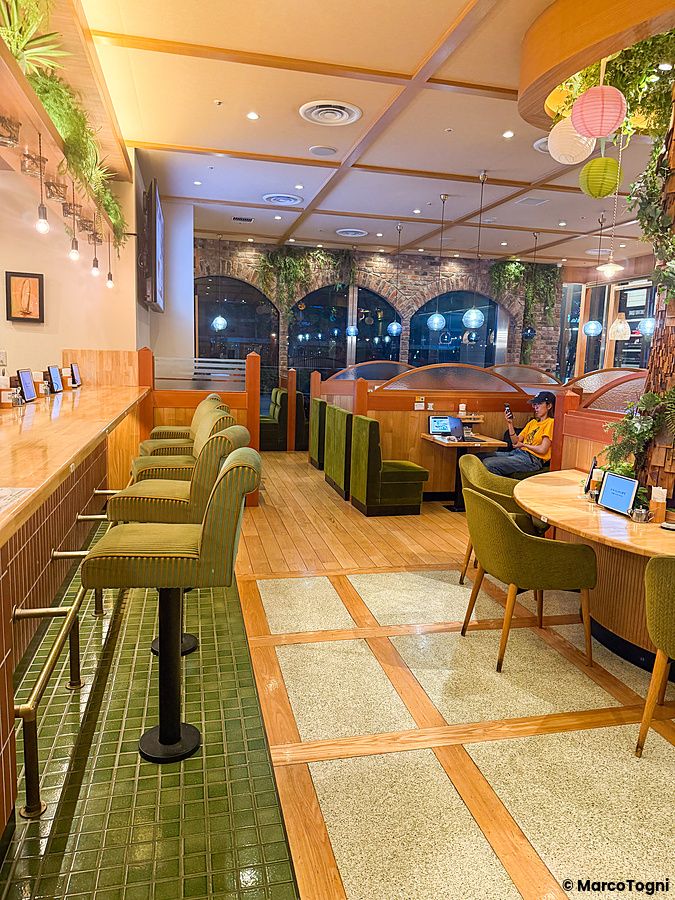 Interno del ristorante vegano Komeda Is a Ginza, con sgabelli verdi, divanetti in legno e una persona che usa un laptop.