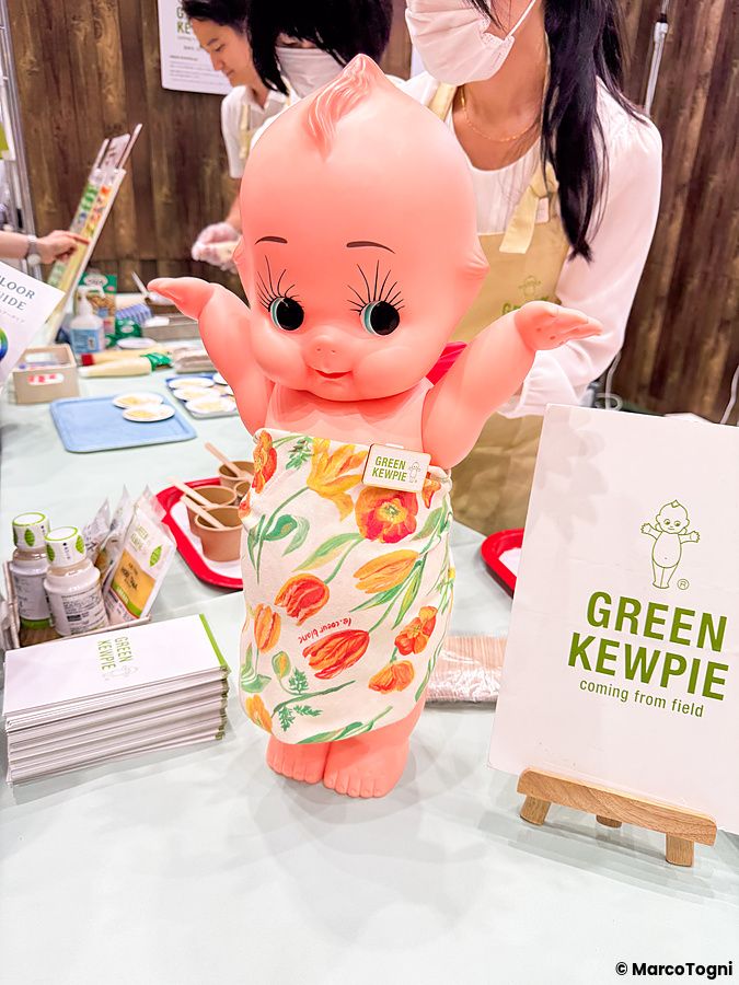 Una statua della mascotte Kewpie su un banco alla Good Life Fair di Tokyo, con dietro persone in uno stand espositivo.