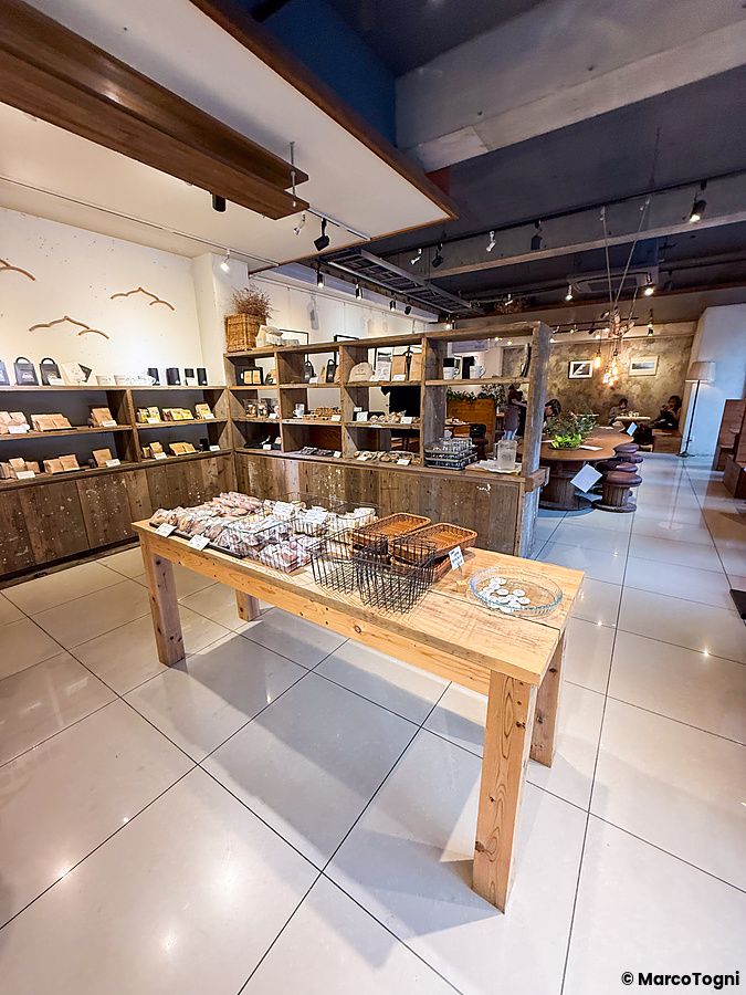 Interno del Takao Coffee con tavoli in legno, scaffalature cariche di prodotti e persone sedute nella zona retrostante.