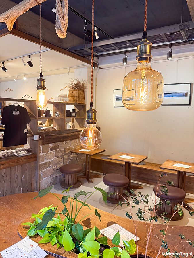 Interno di Takao Coffee con tavoli in legno, sgabelli, lampadine a vista e piante decorative alla base del Monte Takao.
