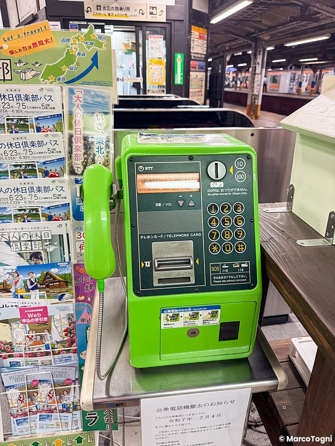 Un telefono pubblico verde NTT situato su una base metallica presso una stazione ferroviaria in Giappone.