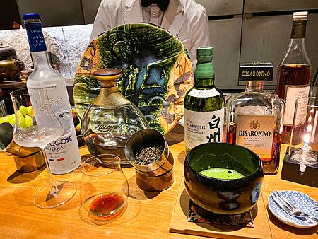 Bottiglie di alcolici, una ciotola di matcha e un piatto decorativo sul bancone in legno del Mixology Salon a Ginza.