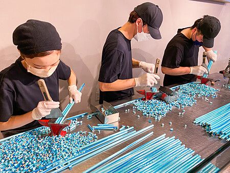 Tre persone con maschere e guanti tagliano lunghi bastoncini di caramelle artigianali azzurre su un tavolo in acciaio.