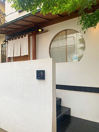 L'ingresso a Sushi Tou mostra una parete bianca con una finestra circolare, un'insegna quadrata e una tenda noren.