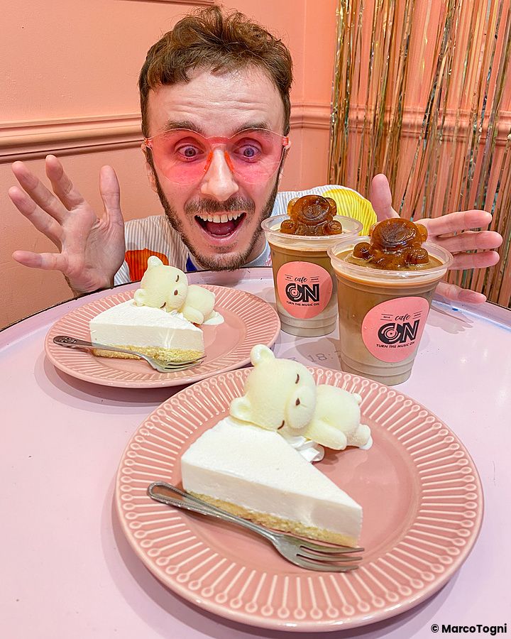 Marco Togni con occhiali rosa siede a un tavolo con dolci e bevande firmate Cafe ON (Shin-Okubo, Tokyo).