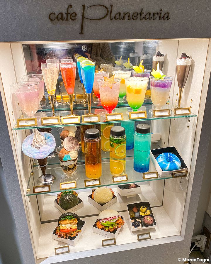 Vetrina del cafe Planetaria a Tokyo con una selezione ordinata di bibite e dolci in esposizione su vari ripiani.