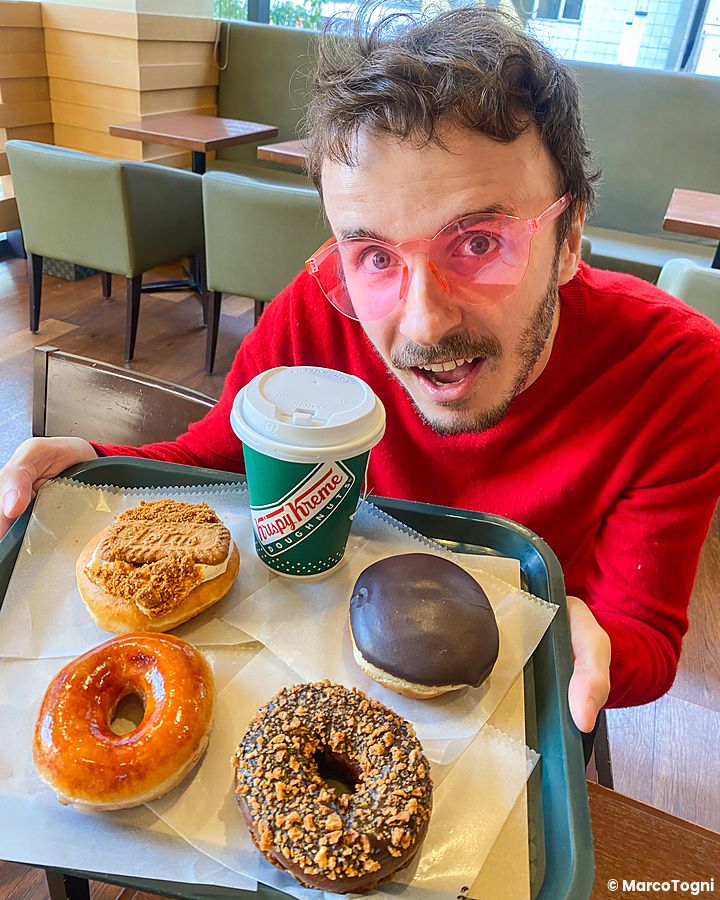 Marco Togni indossa occhiali rosa e mostra un vassoio di ciambelle e un bicchiere Krispy Kreme in un locale moderno.