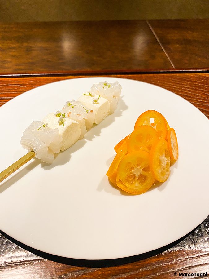 Uno spiedino con formaggio e seppia cruda accompagnato da fette di kinkan su un piatto bianco presso Denkushiflori, Tokyo.