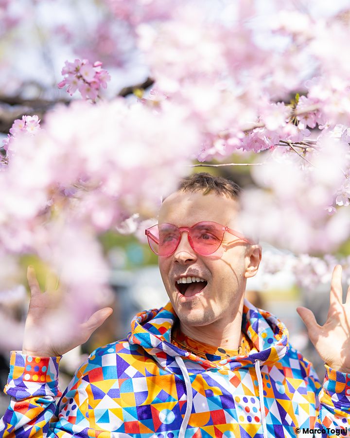 Marco Togni con occhiali rosa e felpa colorata posa tra i fiori di ciliegio in un parco a Tokyo.