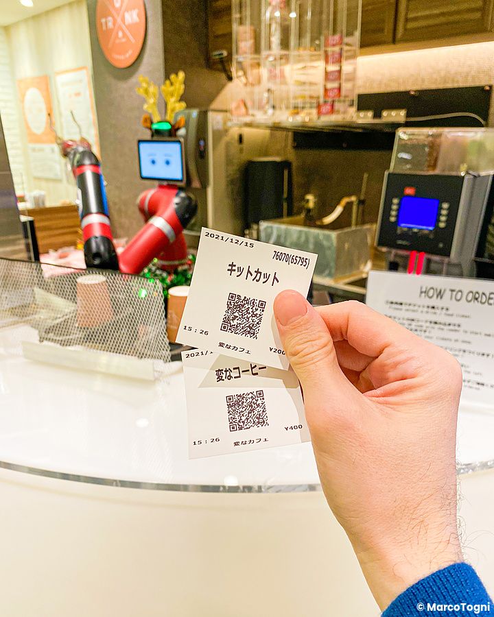 Una mano tiene due scontrini con codici QR davanti a un banco di lavoro con un robot presso l'Henn na Cafe a Shibuya.