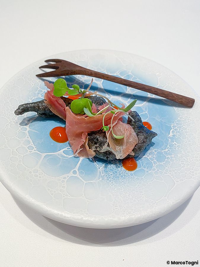 Un antipasto al Koji Shimomura composto da gnocco fritto con calamaro e prosciutto crudo italiano su un piatto azzurro.