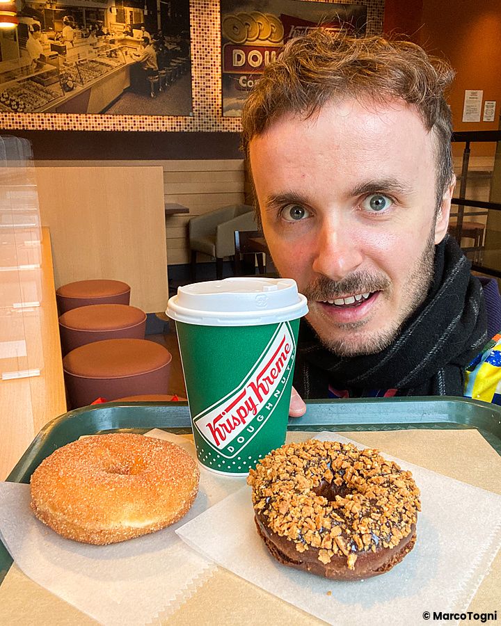 Marco Togni sorridente tiene un vassoio con due ciambelle e un bicchiere di carta brandizzato Krispy Kreme in un locale.