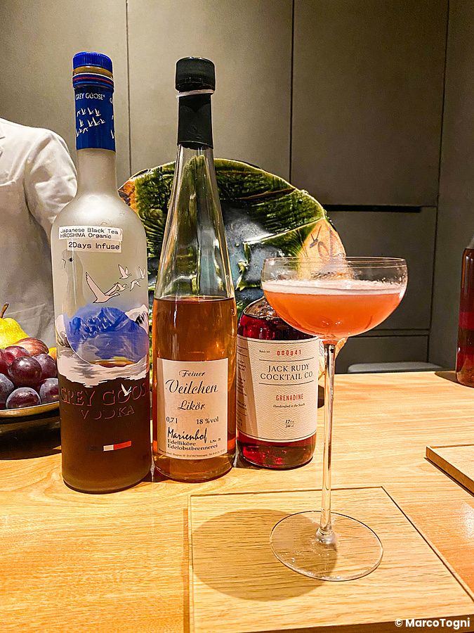 Un cocktail rosa su un bancone di legno al Mixology Salon di Ginza, circondato da bottiglie di distillati e liquori.