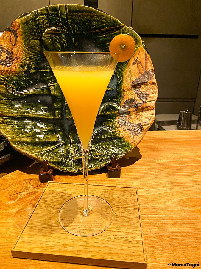 Un cocktail giallo in un calice a V poggia su un vassoio di legno al Mixology Salon di Ginza, con un piatto decorato dietro.