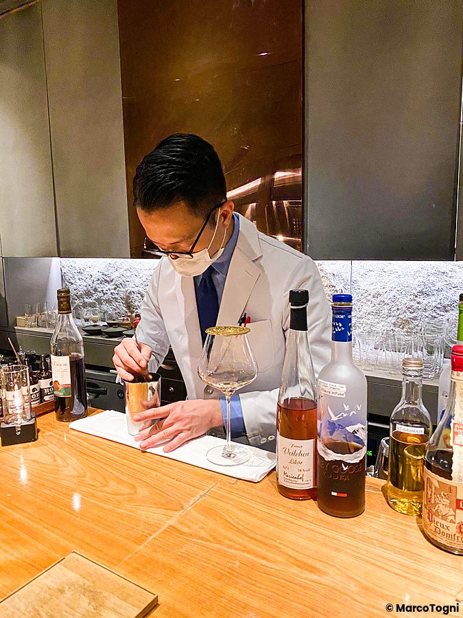 Un barista al Mixology Salon di Ginza prepara un drink dietro un bancone in legno con bottiglie di alcolici.
