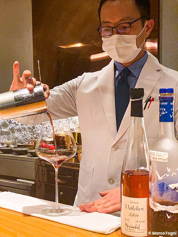Un bartender con camice bianco e mascherina versa un drink in un calice al Mixology Salon di Ginza, Tokyo.