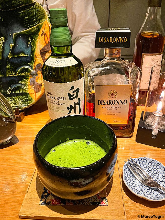 Una ciotola di matcha verde brillante su un bancone di legno accanto a bottiglie di Hakushu e Disaronno al Mixology Salon.