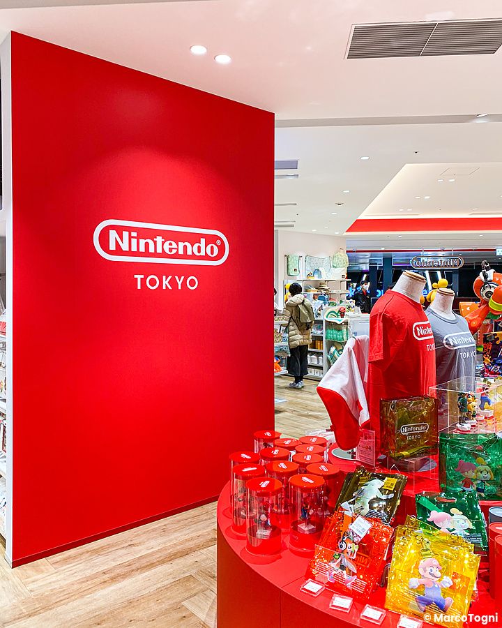 L'interno del Nintendo Store a Tokyo con una parete rossa, logo ufficiale e espositori di merchandising di videogiochi.