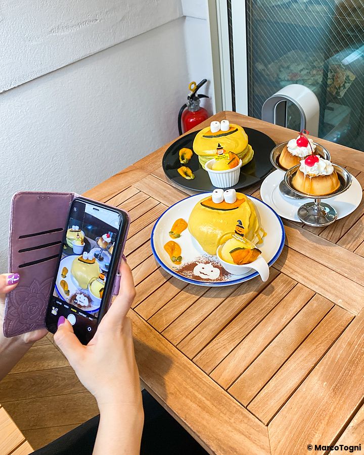 Una persona fotografa con lo smartphone pancake alla zucca a tema Halloween e budini decorati su un tavolo di legno.