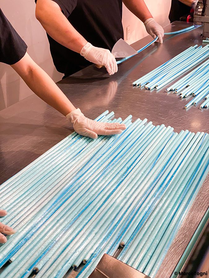 Mani con guanti lavorano lunghi bastoncini di caramelle artigianali azzurre su un tavolo in acciaio al negozio Papabubble.