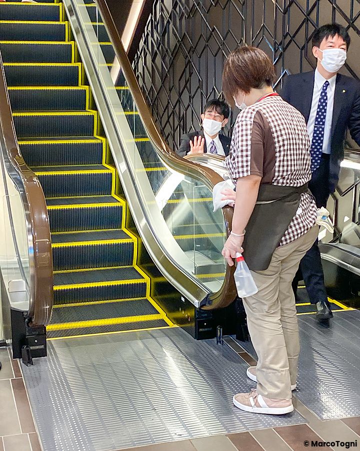 Una persona con divisa a quadri pulisce accanto a una scala mobile a Sapporo, mentre due uomini in abito osservano.