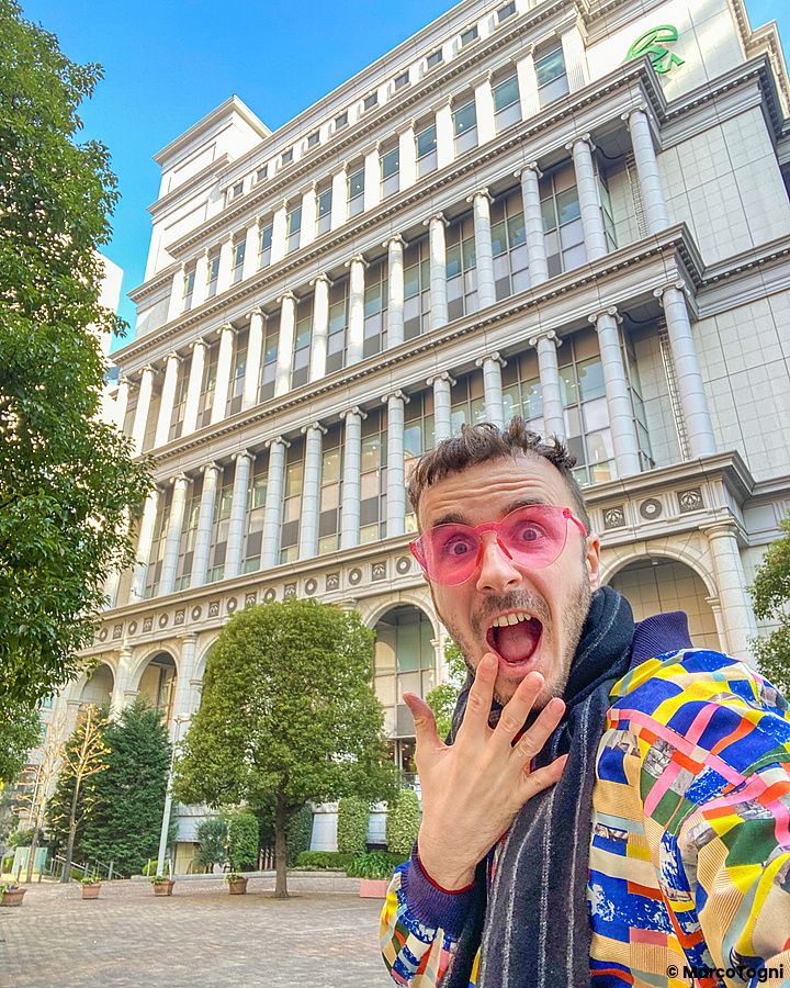 Marco Togni sorridente con occhiali rosa davanti a un edificio nel quartiere italiano a Tokyo.