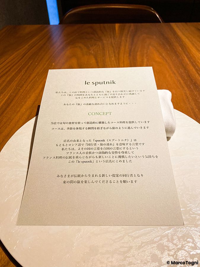 Il menù del ristorante Le Sputnik di Tokyo è appoggiato su un sottopiatto bianco sopra un tavolo in legno.