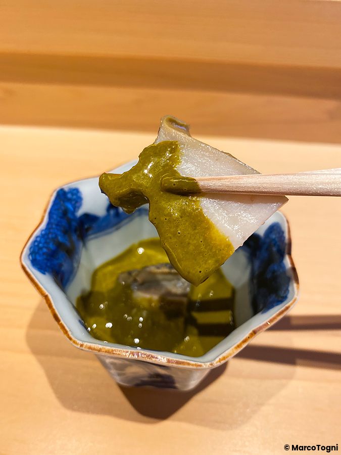 Un pezzo di awabi con salsa fatta con il suo fegato tenuto da bacchette sopra una ciotola da Sushi Masashi.