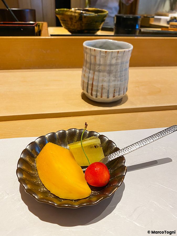 Una porzione di frutta con mango, un pezzo di kiwi e una ciliegia servita su un piccolo piatto da Sushi Takeuchi.