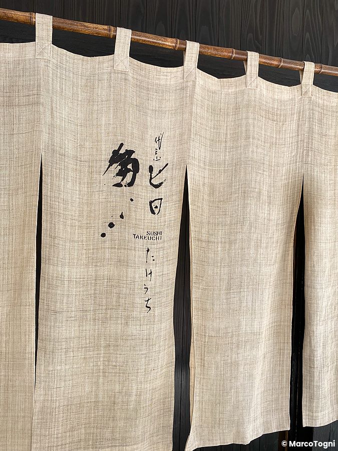L'ingresso a Sushi Takeuchi presenta una tenda noren color beige appesa a un'asta di bambù con scritte in stile calligrafico.