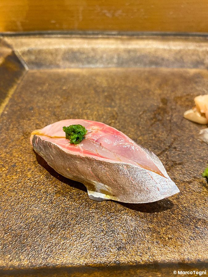 Un nigiri di aji con guarnizione verde servito su un piatto in ceramica scura da Sushi Takeuchi a Tokyo.