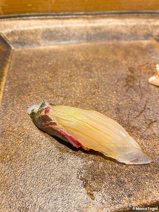 Un singolo nigiri di shima aji posato su un piatto di pietra scura da Sushi Takeuchi.