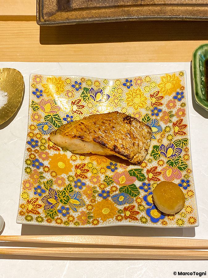 Un filetto di pesce nodoguro grigliato servito su un piatto quadrato con decorazioni floreali a Sushi Takeuchi.