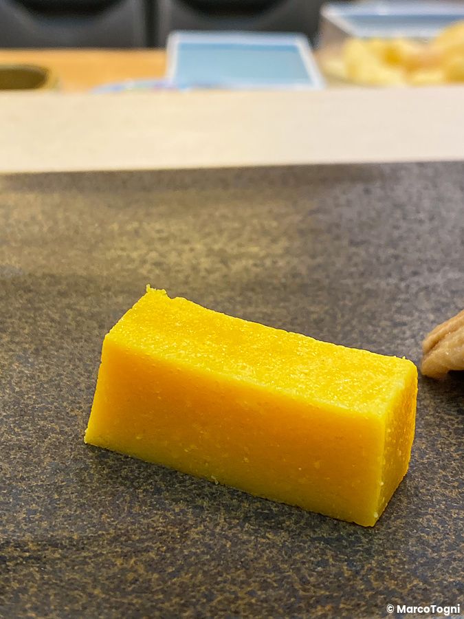 Un pezzo di tamagoyaki rettangolare di colore giallo brillante appoggiato su un piatto scuro da Sushi Tou a Tokyo.