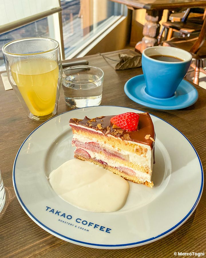 Una fetta di torta alle fragole e cioccolato su un piatto bianco con il logo Takao Coffee accanto a una tazza di caffè.