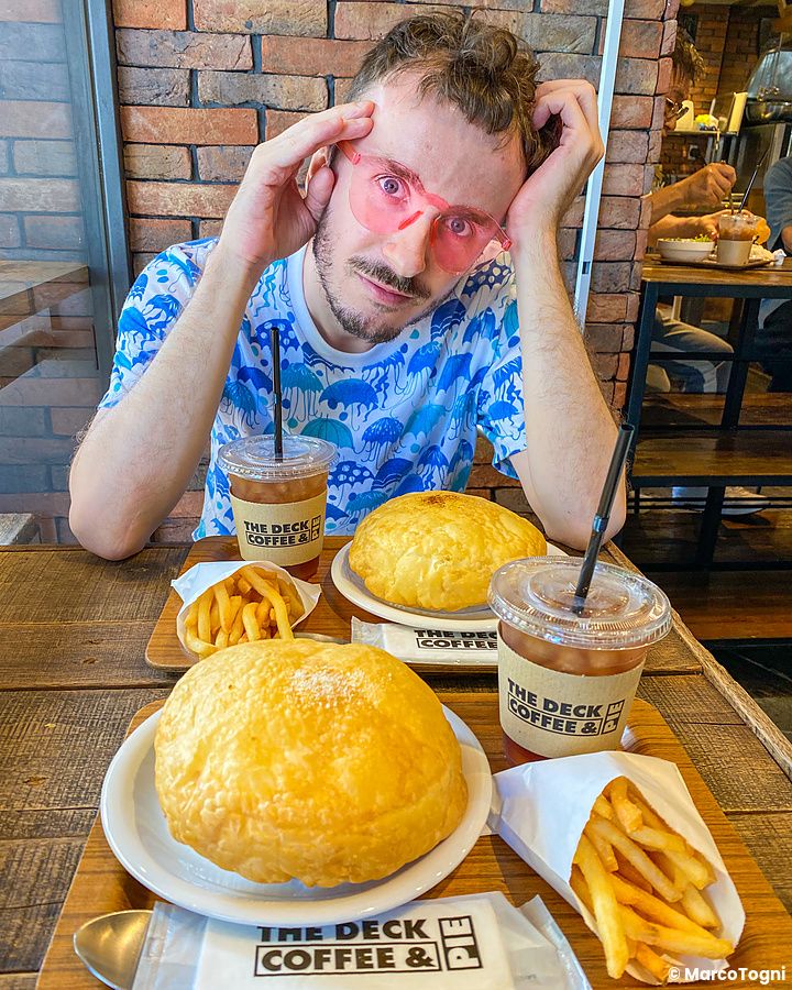 Marco Togni con occhiali rosa siede a un tavolo con hot pie, patatine e bibite al caffè presso The Deck Coffee & Pie.
