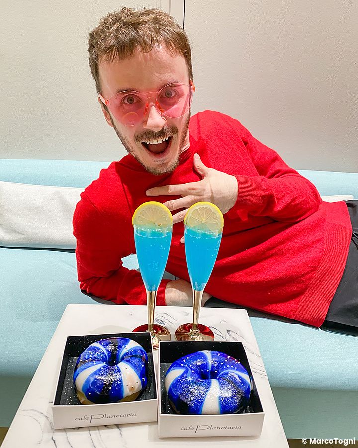 Marco Togni con occhiali rosa nel cafe Tokyo Planetaria con bevande blu e ciambelle decorate in scatole marchiate.