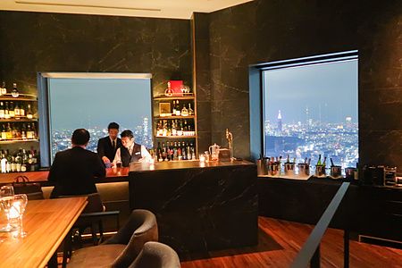 Interno a Bar Bellovisto con vista panoramica notturna su Tokyo attraverso grandi vetrate e persone al bancone.